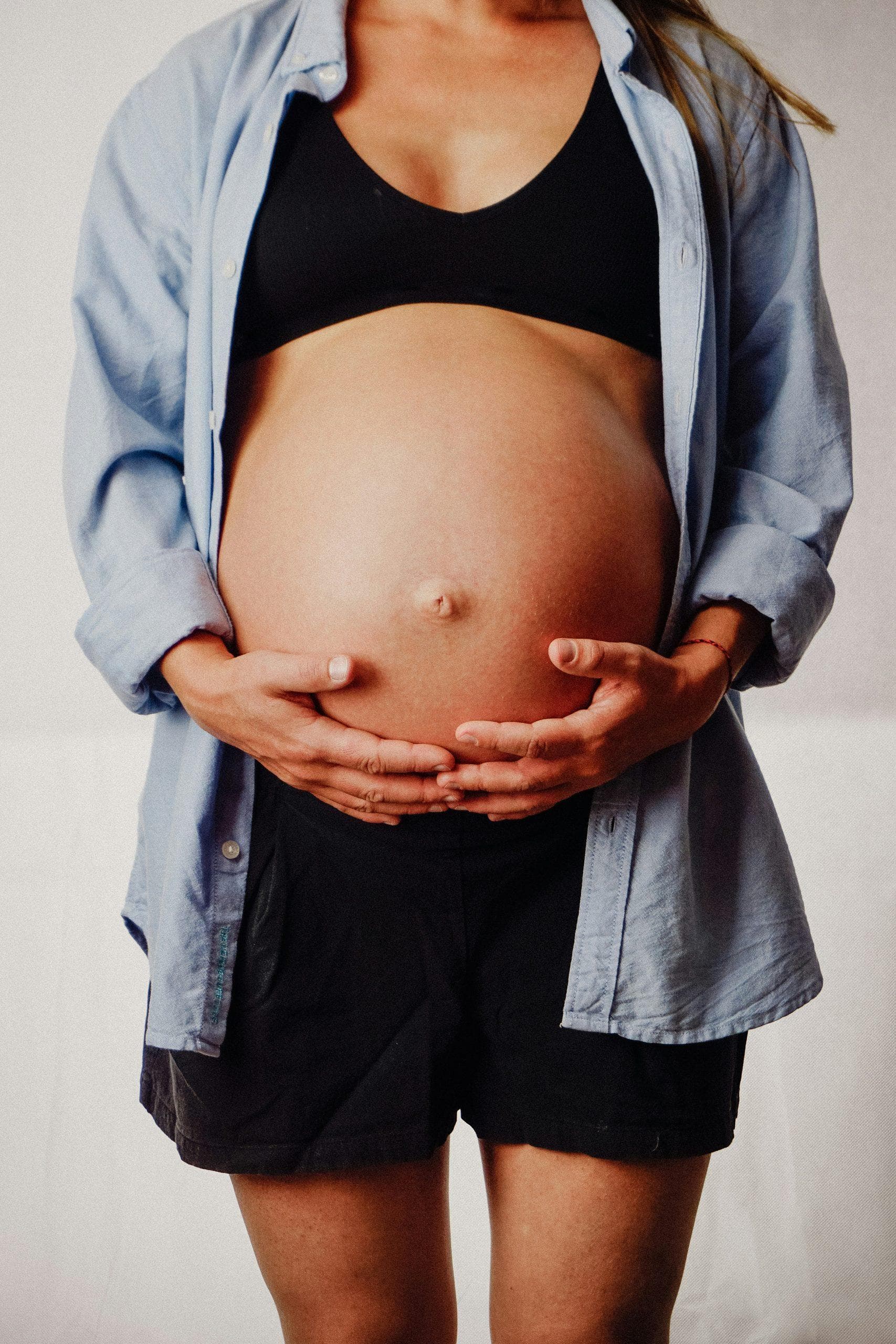A pregnant woman in an open blue shirt, both hands resting under her belly