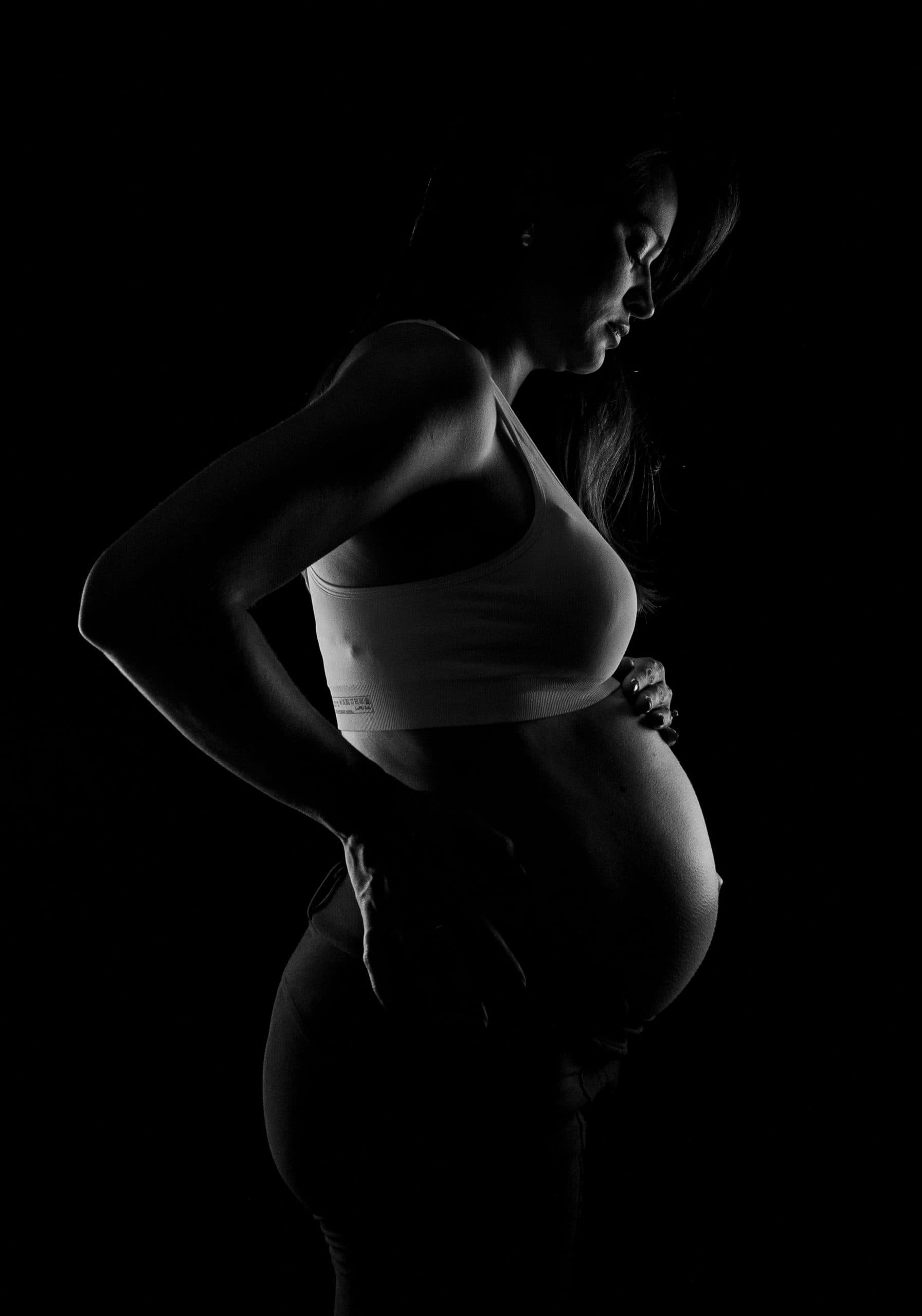 A pregnant woman standing in profile, her outline caught by a single light against the dark