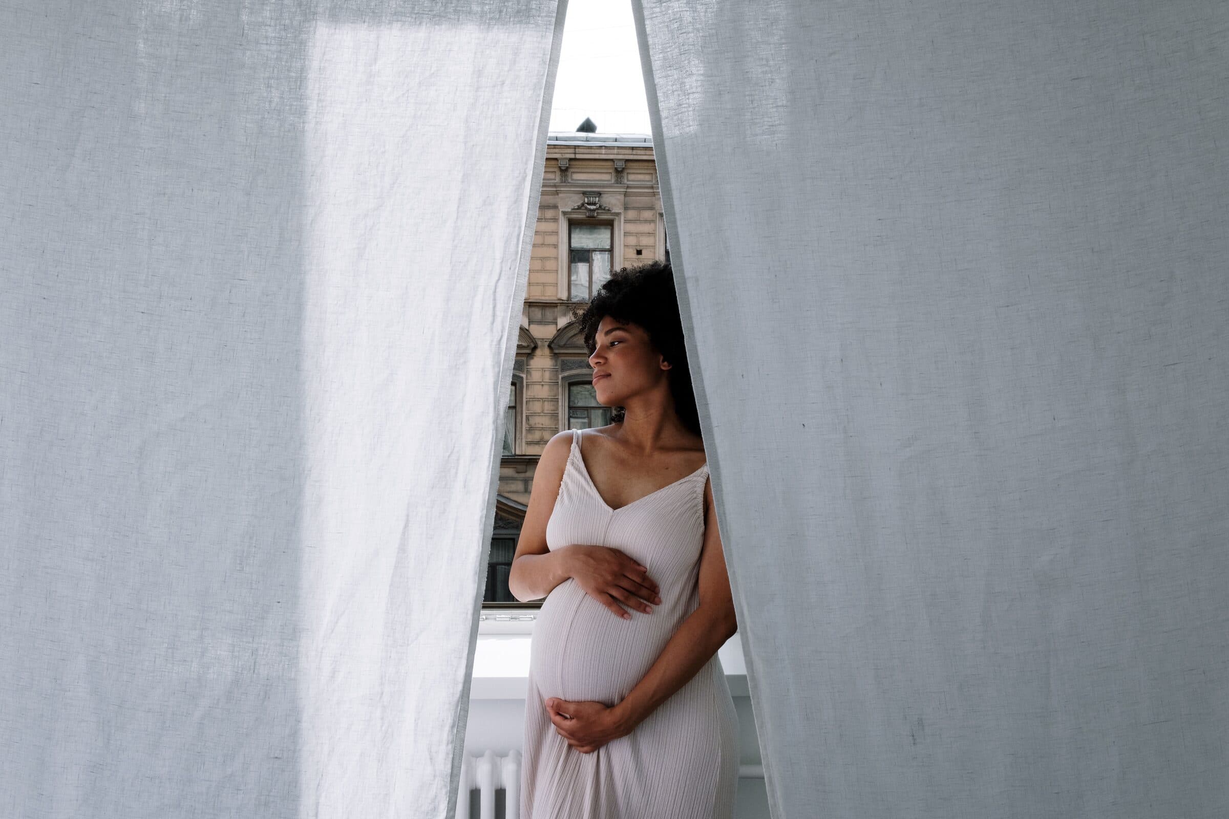 A pregnant woman standing at a window between linen curtains, looking out over the street