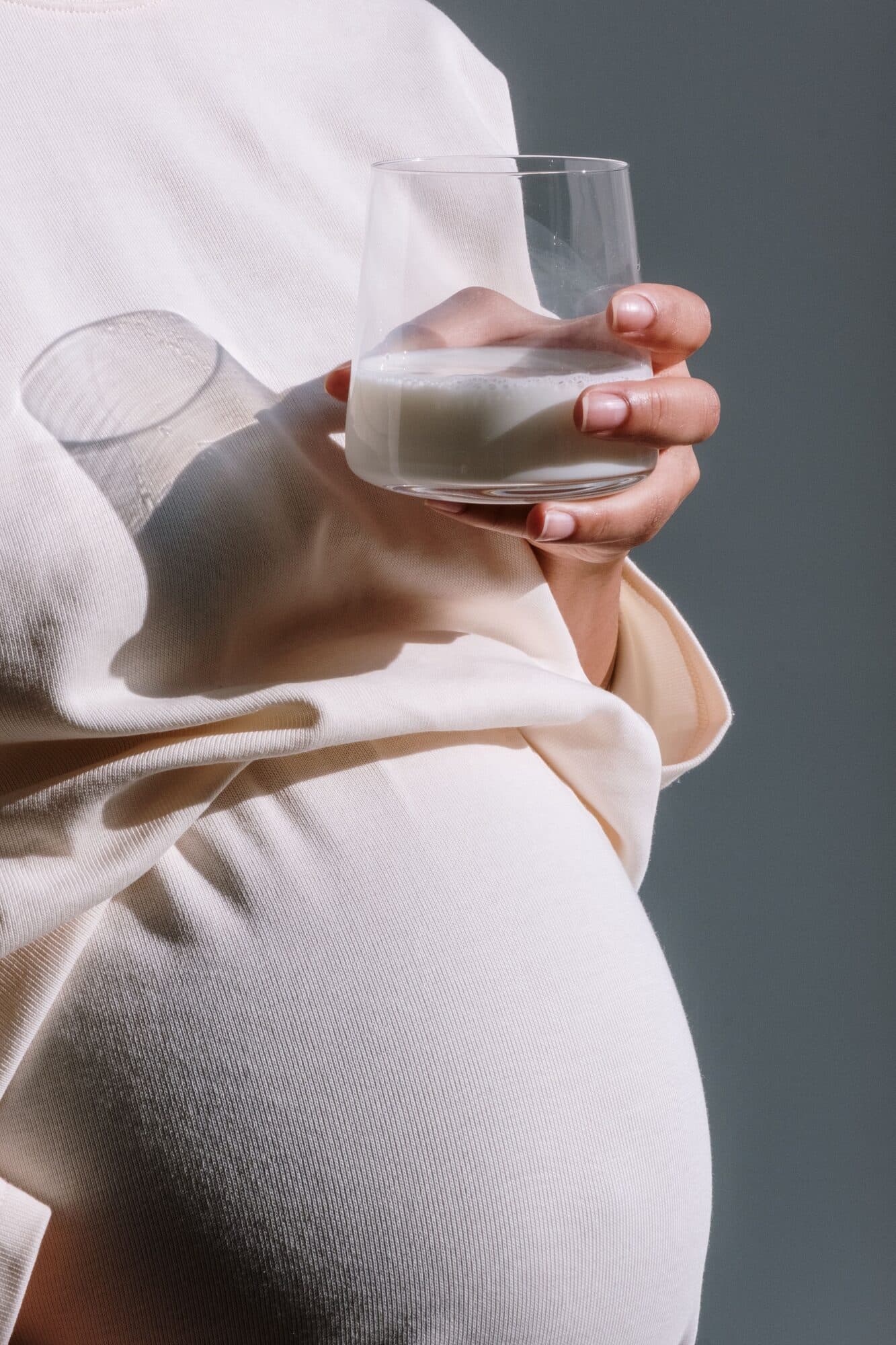 A pregnant woman holding a glass of milk against a soft grey background